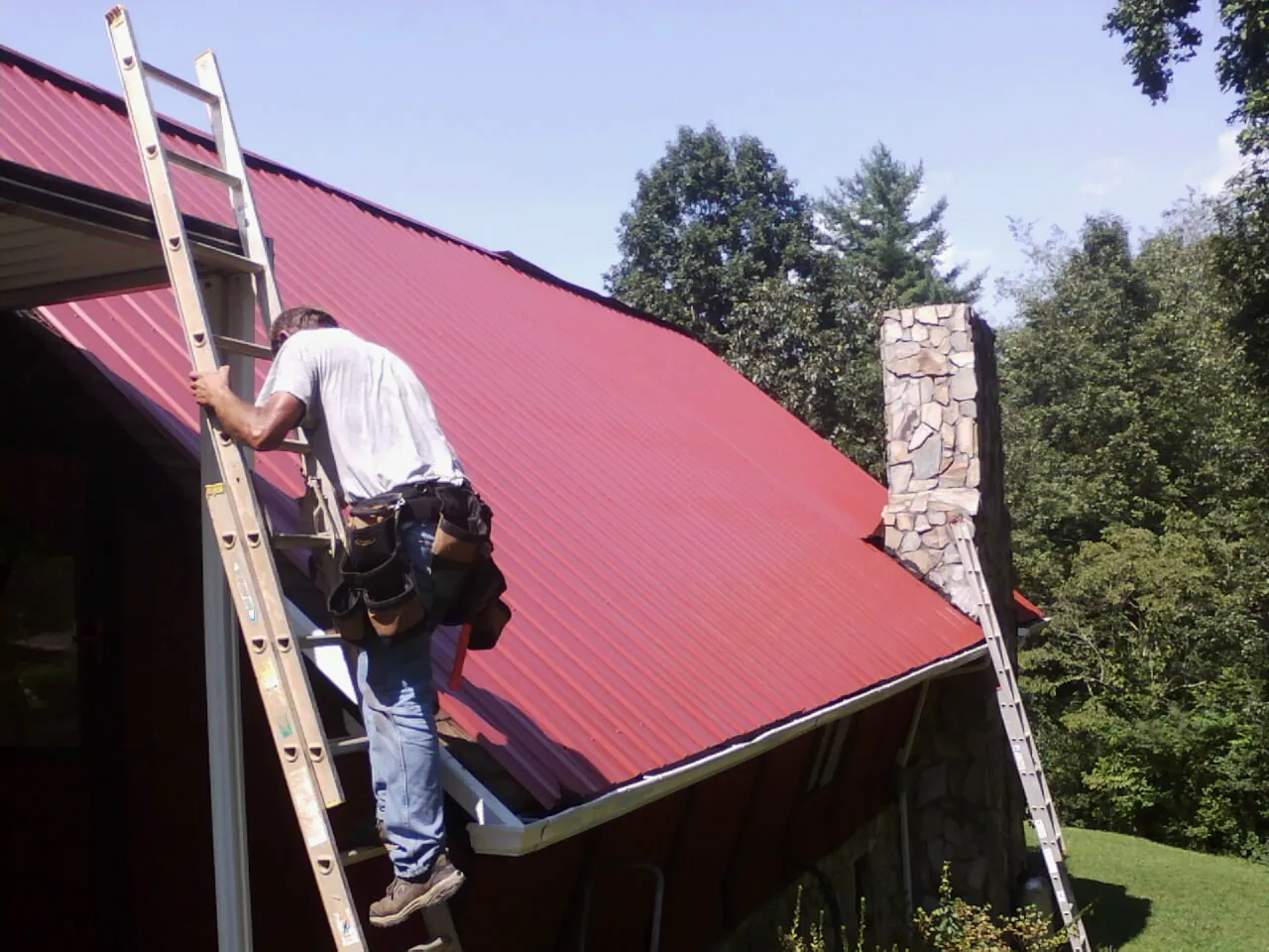 Roofer on a ladder beside a red metal roof and stone chimney — Soffit & Fascia Repair by Roofing Brothers in West Manchester